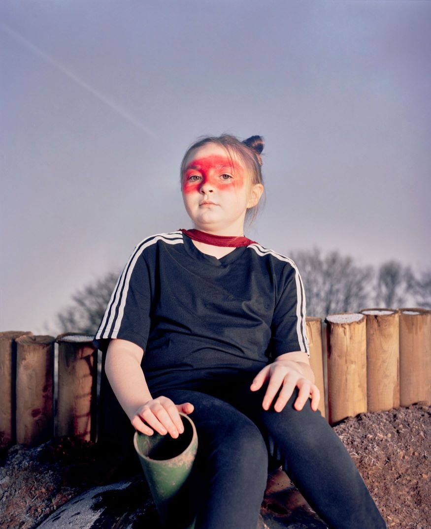 A girl with red make up round her eyes sat in front of a log fence